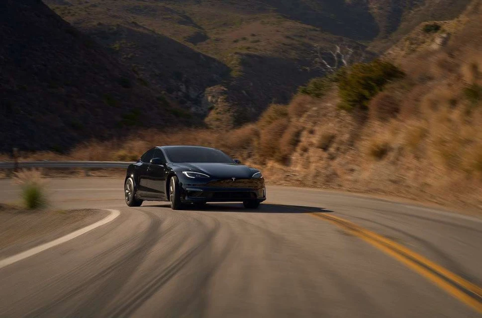 Black electric car drives on asphalt road lined with golden grass, backed by arid hills under a bright sunny sky, in an automotive setting.