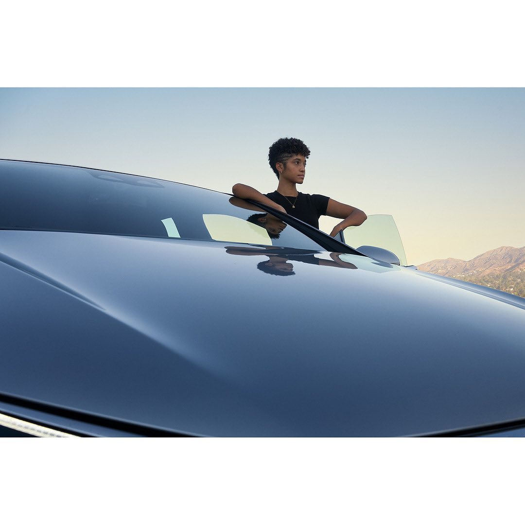 Adult leaning on the hood of a dark blue car with mountains visible in the background under a clear blue sky at dusk.