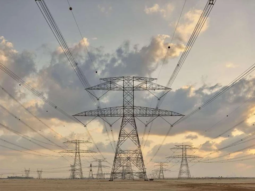 Power transmission towers stand against a cloudy sky, distributing electricity across a flat, sandy landscape at golden hour.