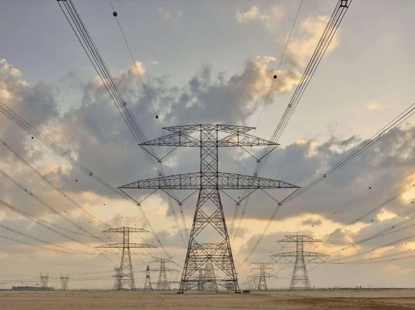 Power transmission towers stand against a cloudy sky, distributing electricity across a flat, sandy landscape at golden hour.