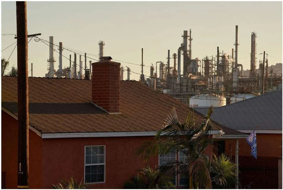 Residential area with single-family homes and a distant industrial oil refinery under a hazy, soft sky in California.