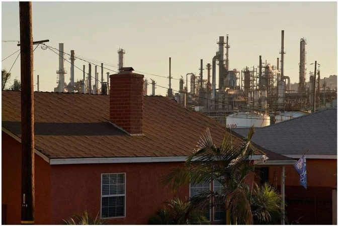 Residential area with single-family homes and a distant industrial oil refinery under a hazy, soft sky in California.