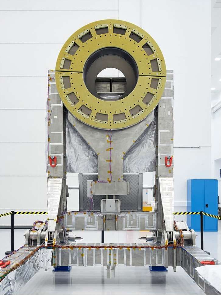 Large piece of industrial equipment with a circular piece on top in an indoor setting, featuring metallic surfaces, a yellow frame, and blue cabinet.