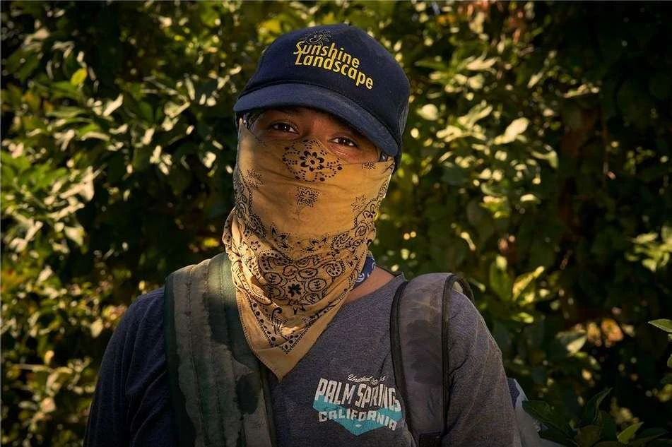 A person wearing a hat and bandana stands outside surrounded by green leaves in natural light with dark shadows and a backpack.