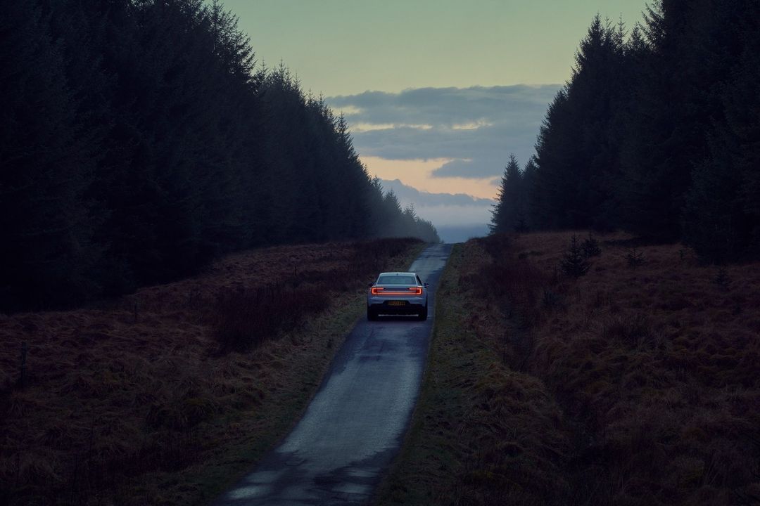Electric car driving on a dark rural road lined with tall, dark trees under a cloudy sky at dusk, automobile tail lights illuminated.