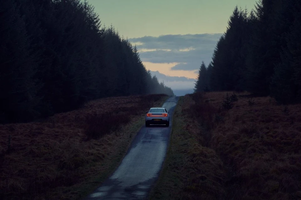 Electric car driving on a dark rural road lined with tall, dark trees under a cloudy sky at dusk, automobile tail lights illuminated.