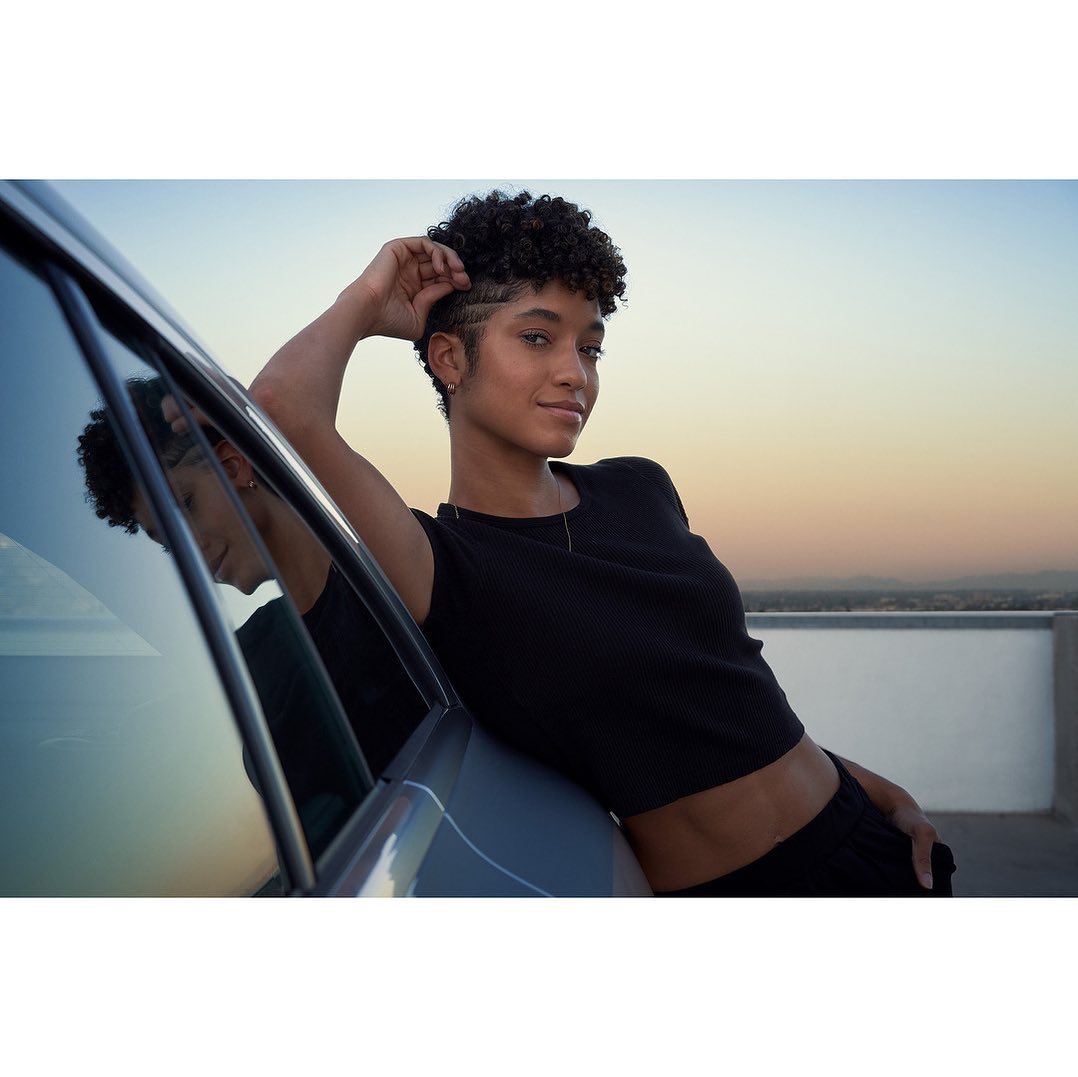 A woman with an Afro and a black top stands next to a car on a rooftop against a gradient sunset, smiling at the viewer.