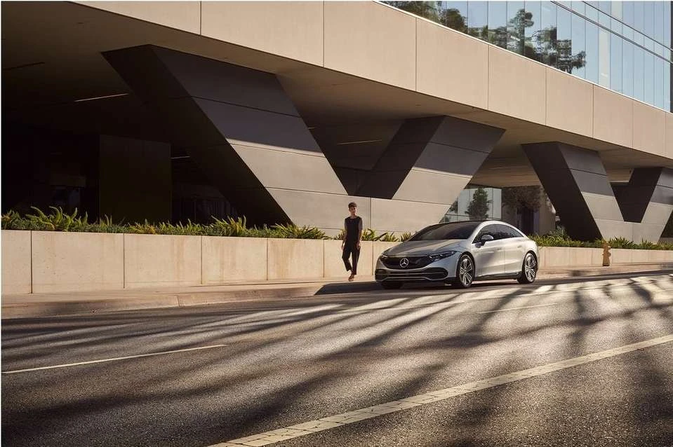 Silver Mercedes-Benz EQS parked on a city street with a person walking by a modern building with geometric architectural details.