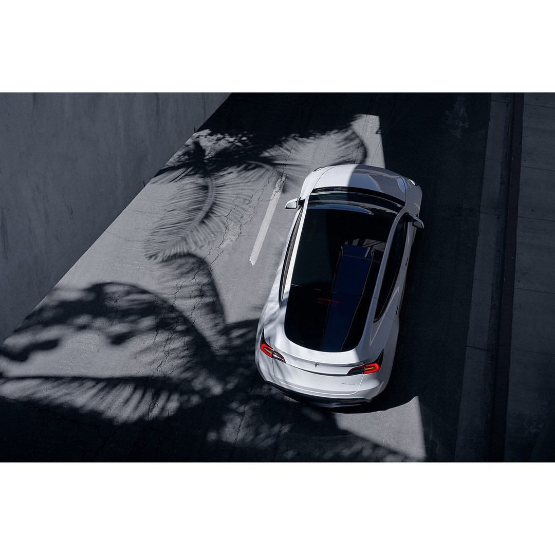 Aerial view of a white car parked on asphalt with leaf shadows creating texture, next to a light gray wall, showcasing automotive design.