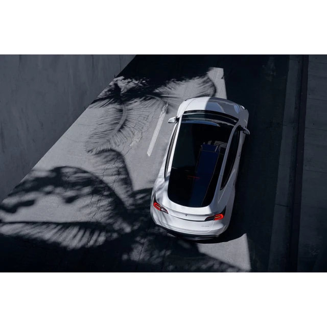 Aerial view of a white car parked on asphalt with leaf shadows creating texture, next to a light gray wall, showcasing automotive design.