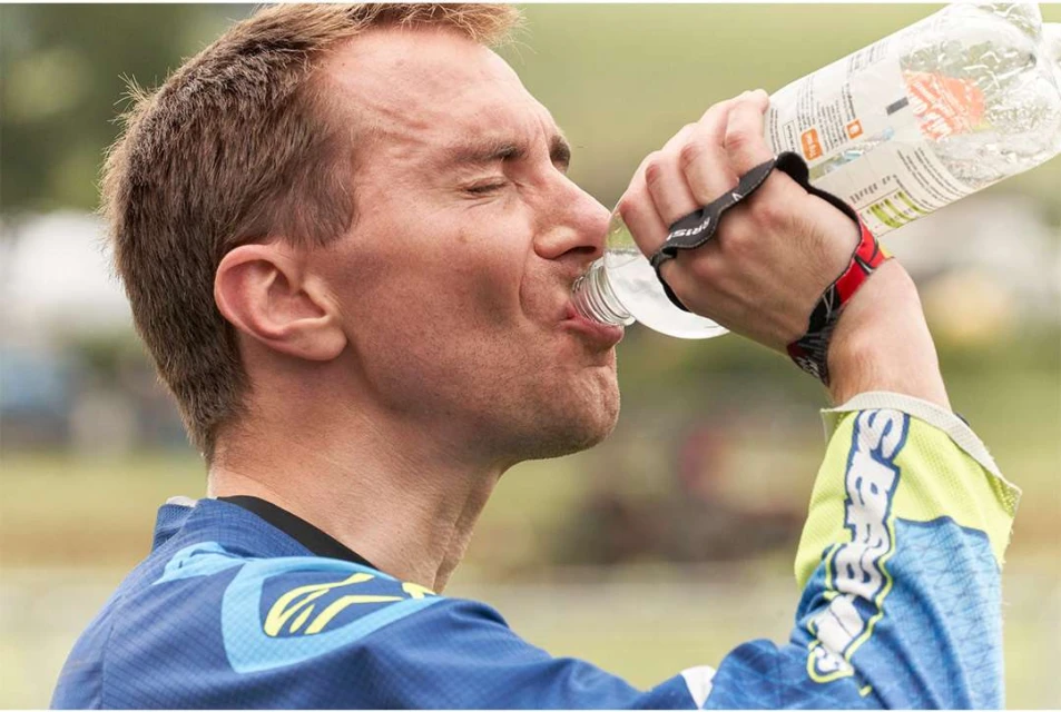 Adult male athlete drinking from a water bottle with greenery in the background, showcasing hydration and outdoor fitness.