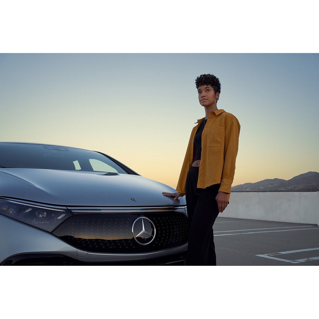 An adult woman in a yellow shirt stands next to a silver electric car on a rooftop parking lot with a mountain range in the distance.