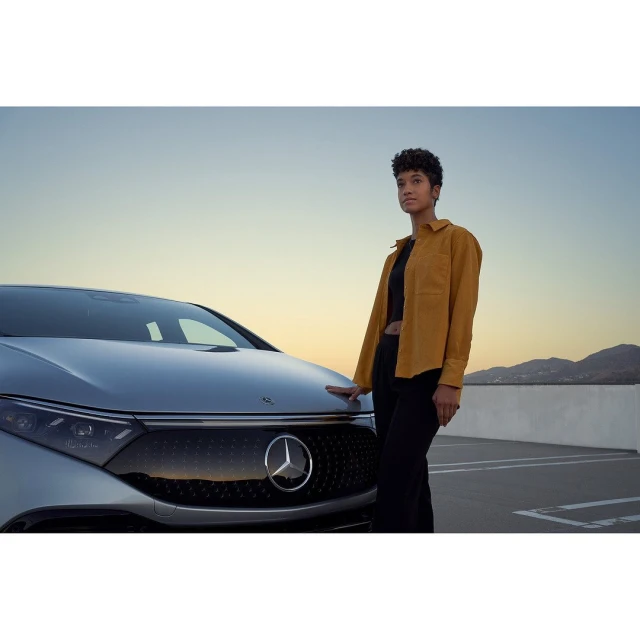 An adult woman in a yellow shirt stands next to a silver electric car on a rooftop parking lot with a mountain range in the distance.