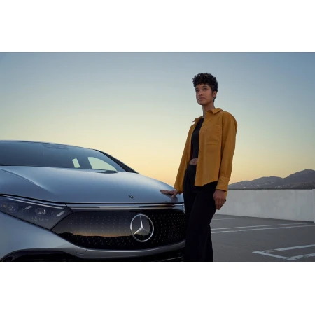 An adult woman in a yellow shirt stands next to a silver electric car on a rooftop parking lot with a mountain range in the distance.