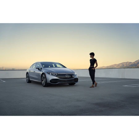 Silver Mercedes-Benz EQS parked on concrete with woman standing next to it in outdoor setting at daytime with clear sky.