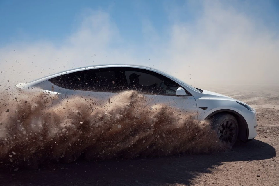 A white Tesla car kicks up dirt while driving on a dry, dusty road under a bright, cloudless sky, showcasing automotive performance.