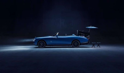 Side view of a luxury blue convertible car parked in a dark studio with its trunk open, featuring a matching umbrella and two folding stools