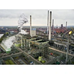 Aerial view of a sprawling industrial factory complex with tall smokestacks billowing white smoke and complex piping networks.