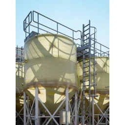 Large industrial storage tanks with steel platforms and safety ladders under a clear blue sky, photographed by Greg White.