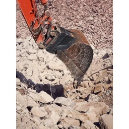 Close-up of an orange excavator bucket positioned above broken concrete rubble and debris at a construction site