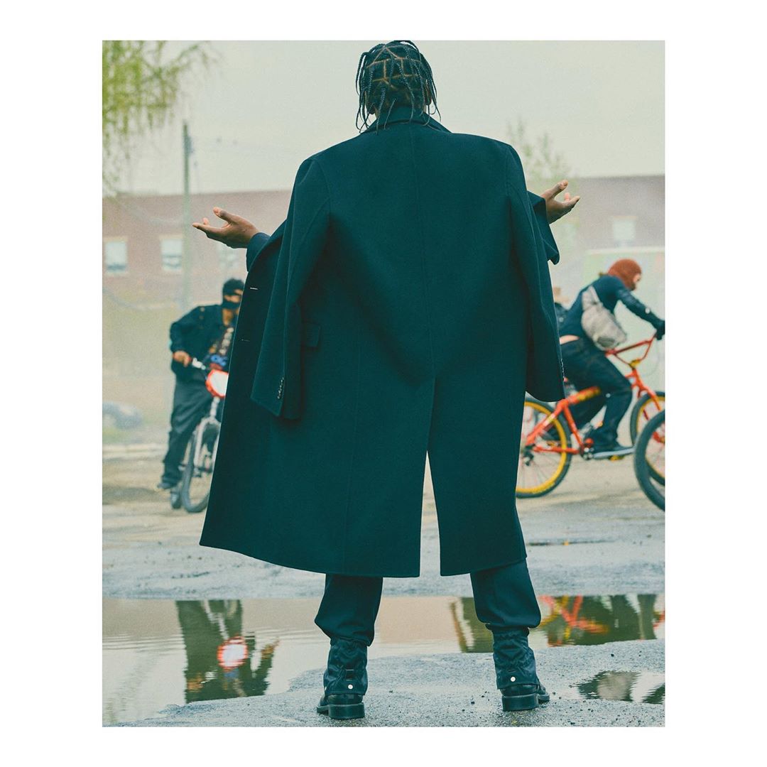 Man in oversized black coat with arms outstretched, standing in puddle with bicycles riding in background, urban gritty scene.