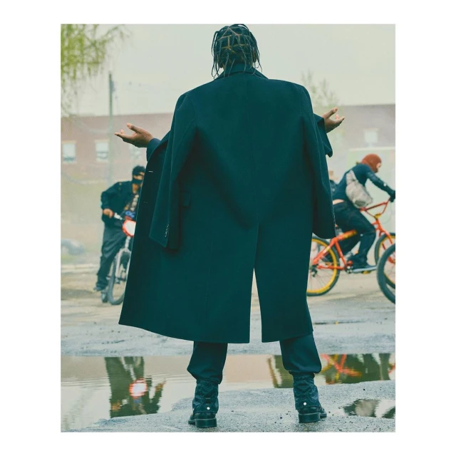 Man in oversized black coat with arms outstretched, standing in puddle with bicycles riding in background, urban gritty scene.