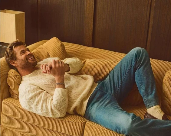 Chris Hemsworth smiling while lying on a ribbed mustard yellow corduroy couch wearing a cream cable knit sweater and blue jeans