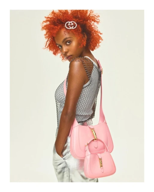 Fashion editorial shot of a woman with orange hair wearing a grey top and holding two pink purses, against a light neutral backdrop.