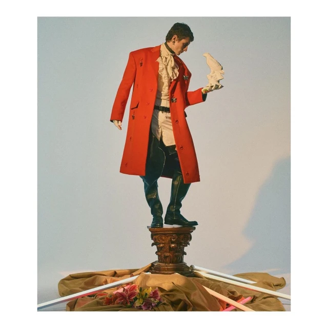 Man stands on pedestal in red coat holding bird statue.  He wears black boots, white pants and frilly shirt. Studio lighting, light background.
