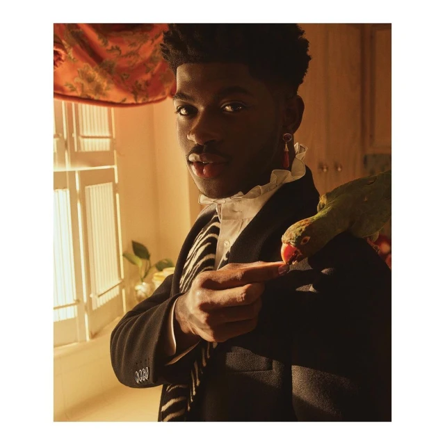 Lil Nas X poses indoors with a green parrot perched on his shoulder, wearing a black coat, white shirt, and zebra-striped scarf.