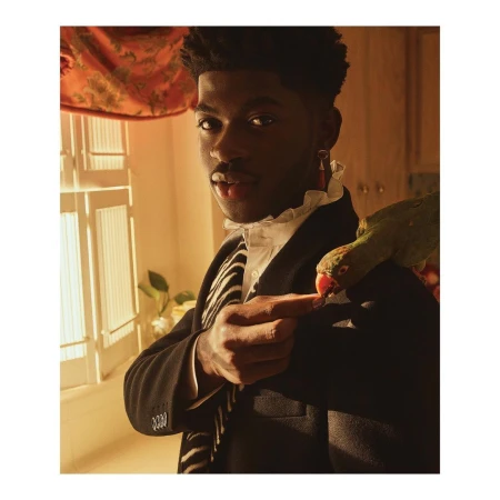 Lil Nas X poses indoors with a green parrot perched on his shoulder, wearing a black coat, white shirt, and zebra-striped scarf.