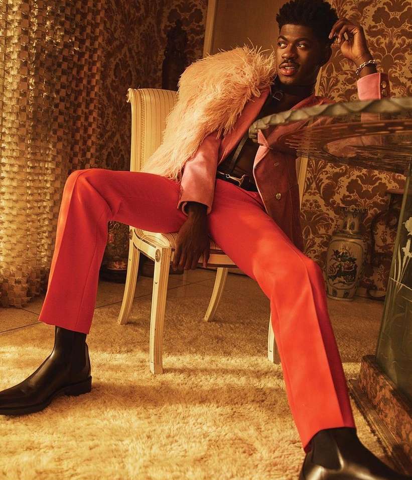 A stylish adult male sits in an ornate room wearing a peach-colored suit with a feathered boa and black boots, gazing at the camera.