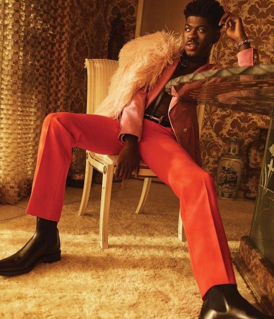 A stylish adult male sits in an ornate room wearing a peach-colored suit with a feathered boa and black boots, gazing at the camera.