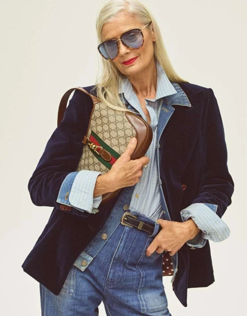 Older female model in a blue denim outfit and navy velvet blazer holds a Gucci handbag against a plain background in a studio shot.