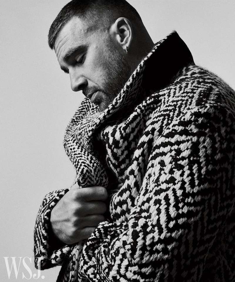 Portrait of a man in a patterned coat looking down, captured in black and white. The subject is well-groomed with short, styled hair.