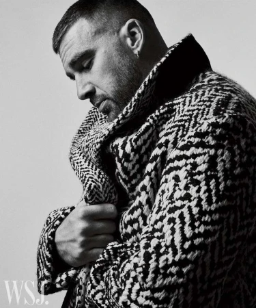 Portrait of a man in a patterned coat looking down, captured in black and white. The subject is well-groomed with short, styled hair.