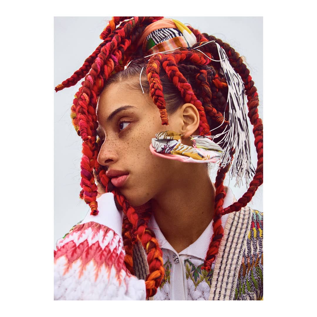 Close-up of a woman with freckles and colorful braided hair wearing patterned clothing and large earrings against a pale background.