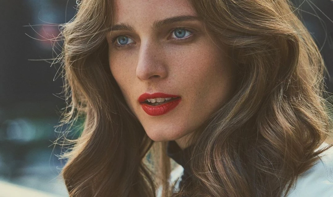 Close-up portrait of a woman with wavy brown hair, bright blue eyes, and red lipstick, set against a blurred dark background.