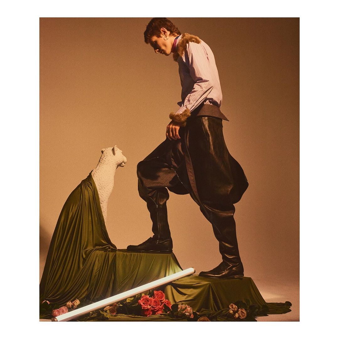 Fashion shot of a young adult male wearing a lilac shirt and baggy black pants standing on a platform next to a white statue.