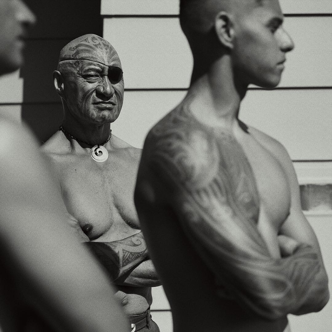 Three men are standing outdoors in black and white. One man has face tattoos and an eye patch, and the other two have tattoos on their arms.