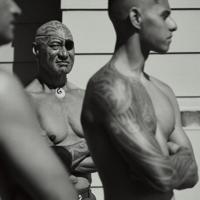 Three men are standing outdoors in black and white. One man has face tattoos and an eye patch, and the other two have tattoos on their arms.