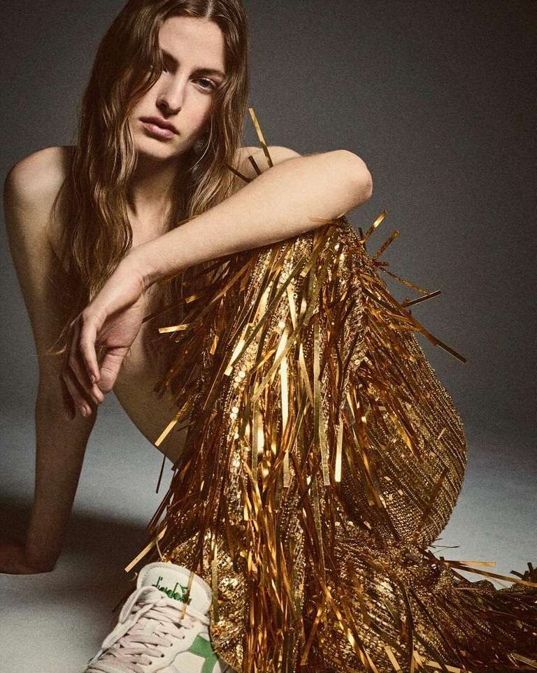 Adult woman poses in a studio, wearing a gold fringe skirt and white sneakers, exhibiting fashion and style against a grey backdrop.
