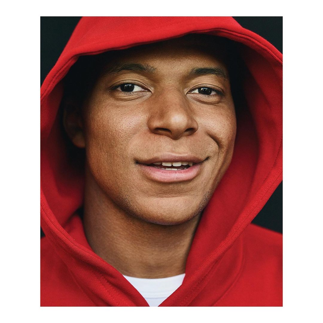 Close-up of a smiling adult male with dark hair wearing a red hooded sweatshirt against a dark background in a portrait orientation.
