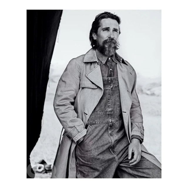 Black and white portrait of a bearded man wearing a denim jumpsuit and trench coat, posing outdoors with a neutral expression and gazing off-camera.