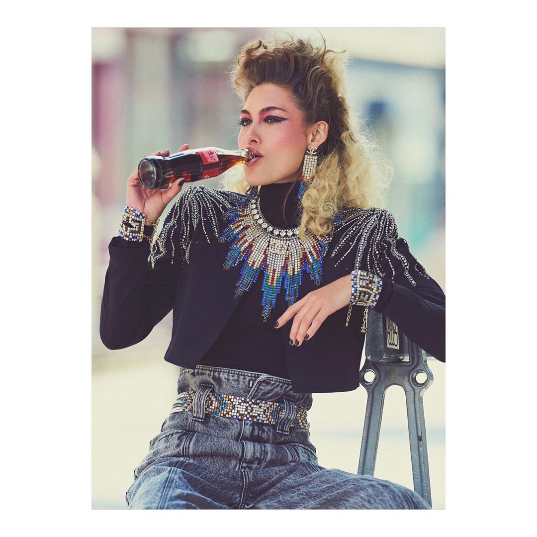 Adult woman with wavy blonde hair drinks from a Coca-Cola bottle while seated, wearing a beaded black jacket and acid wash jeans.