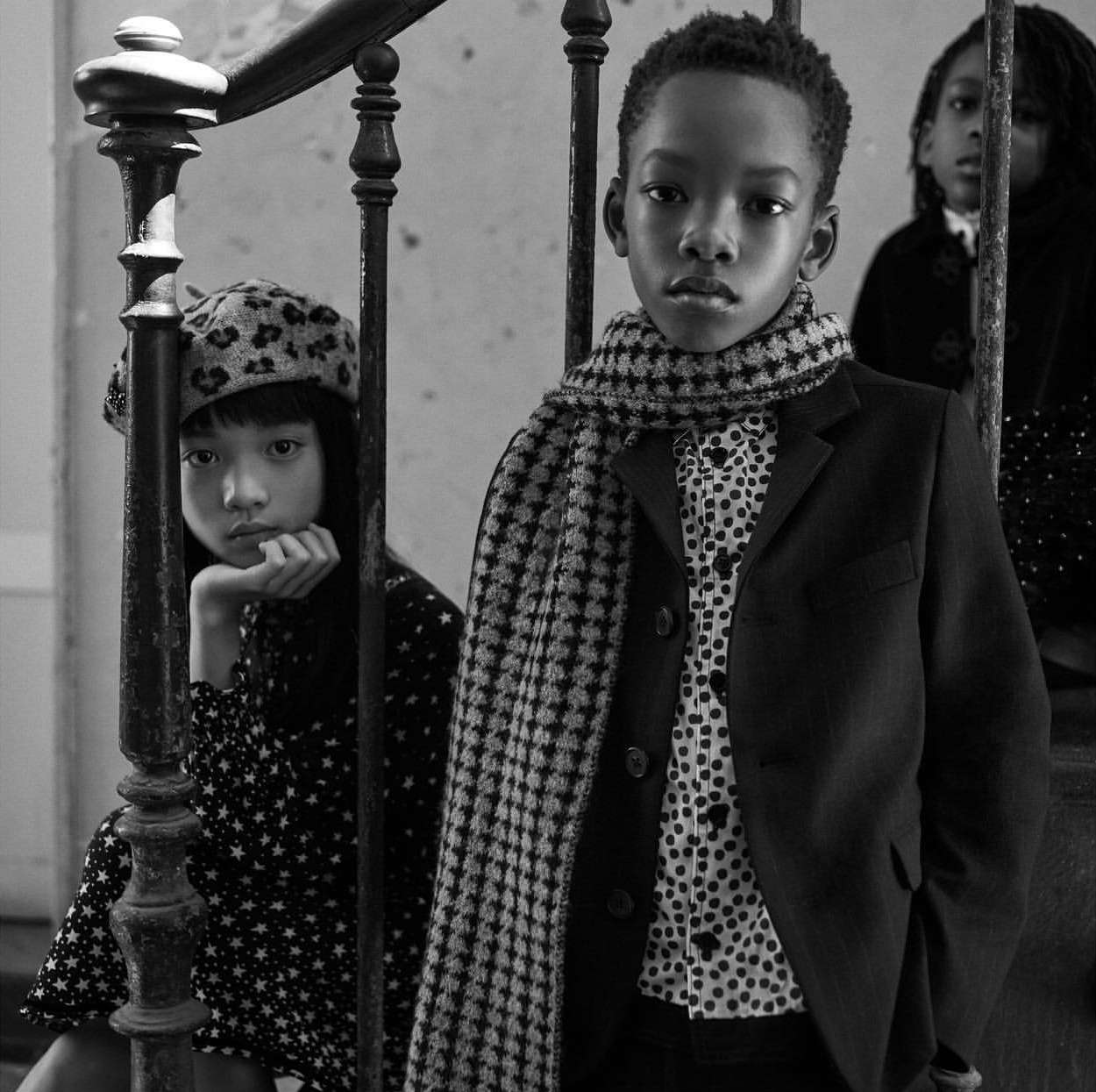 Black and white shot of a young Black boy wearing a suit and scarf standing next to a young Asian girl in a patterned dress.