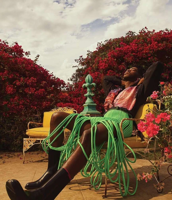 Person relaxes in an outdoor chair surrounded by bright bougainvillea. They wear a neon green rope skirt, black boots, and a patterned shirt.