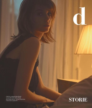 A woman in a black lace top seated indoors under warm interior lighting with a bedside lamp and draped curtains.