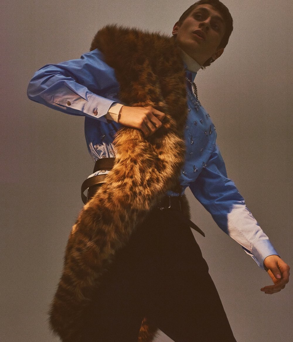 A stylish adult male poses in a studio wearing a blue shirt with embellishments, dark trousers, and a spotted fur stole.