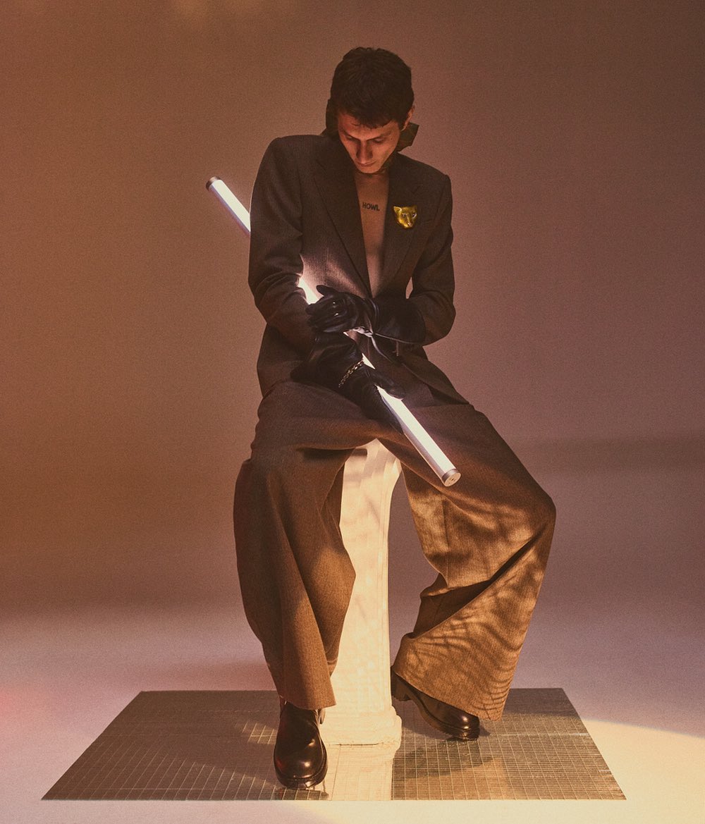 An adult male sits in a studio wearing a suit with wide-leg pants, black leather boots, and gloves, holding a light stick.